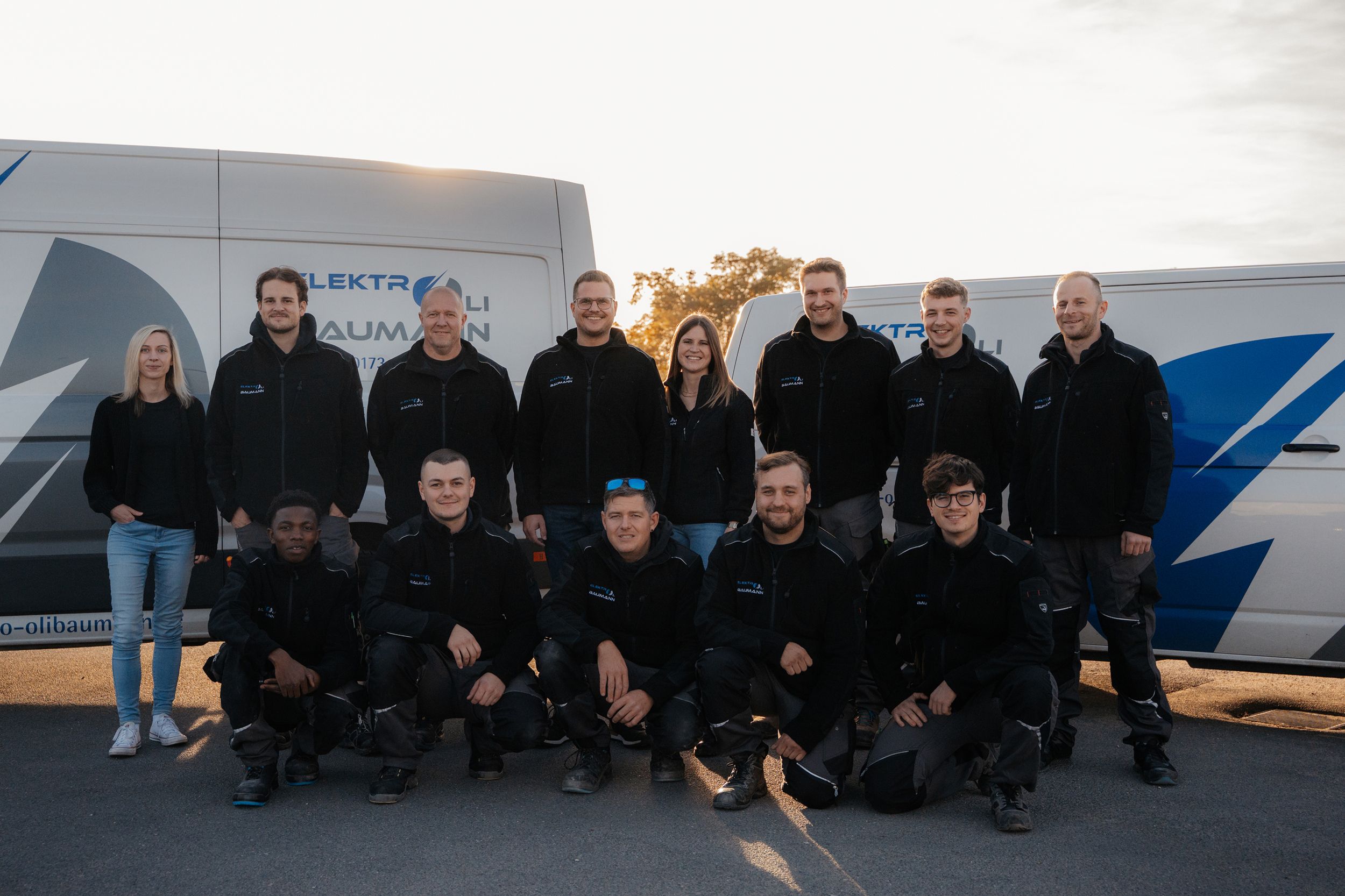 Ein Gruppenfoto des gesamten Firmenteams in einheitlicher dunkler Arbeitskleidung, das vor zwei weißen Firmenfahrzeugen mit Logo im warmen Licht der tiefstehenden Sonne posiert